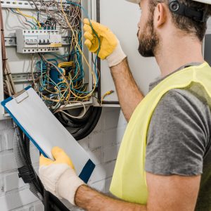 electricista sosteniendo portapapeles y revisando cables en caja eléctrica en corredor